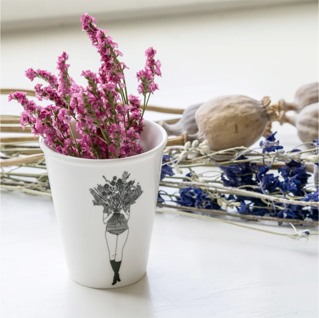 taza de porcelana con el dibujo en negro de una chica con la cara tapada por un ramo de flores y unas flores en la taza