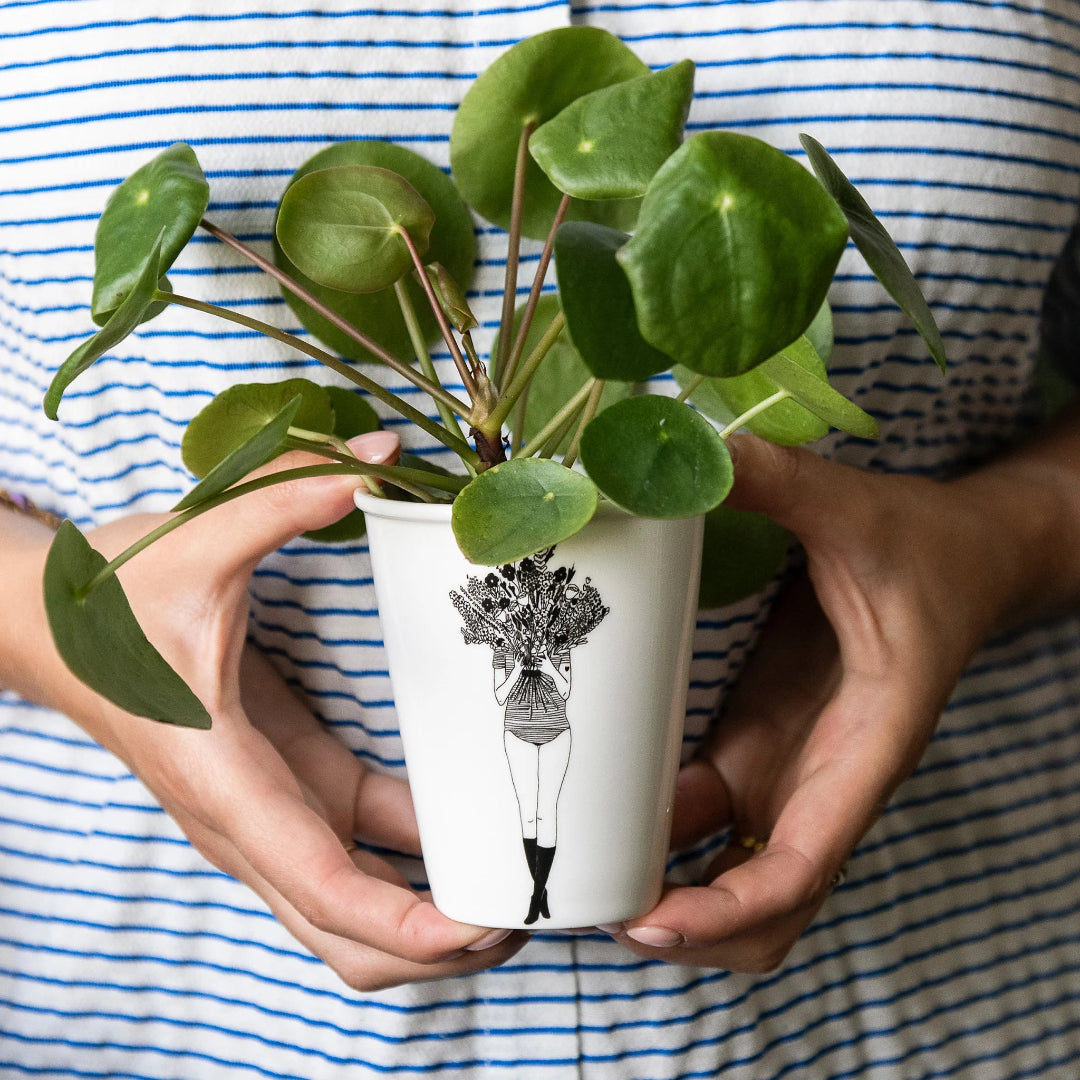 taza de porcelana con el dibujo en negro de una chica con la cara tapada por un ramo de flores y una planta dentro de la taza