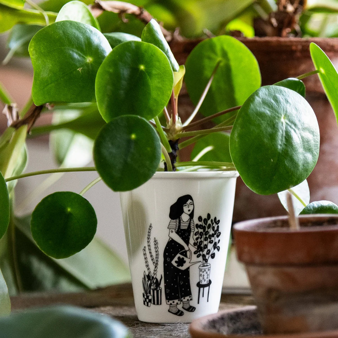 Vaso de cerámica con ilustración de una chica vestida con un mono regando las plantas usado como macetita en un jardín con una pilea sembrada