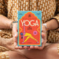Mujer sentada en postura de loto sosteniendo una guía con cartas ilustradas de diferentes posturas de yoga y manual de uso