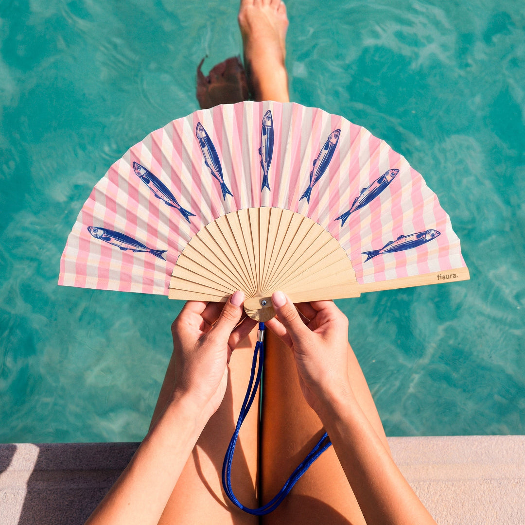 persona en una piscina posando con el abanico de la marca fisura con el fondo a rayas verticales rosas y blancas y con estampado de sardinas en color azul oscuro, abierto y con la cuerda en color azul oscuro para engancharla 