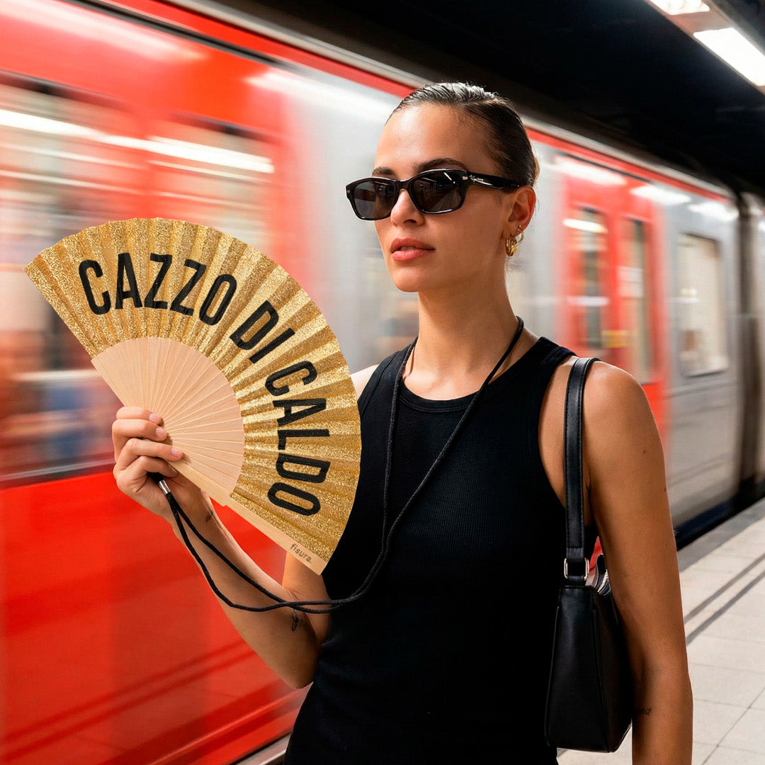 chica sosteniendo el abanico de la marca fisura de tela y madera en color purpurina dorada con el texto CAZZO DI CALDO en negro, mientras un metro pasa por detrás