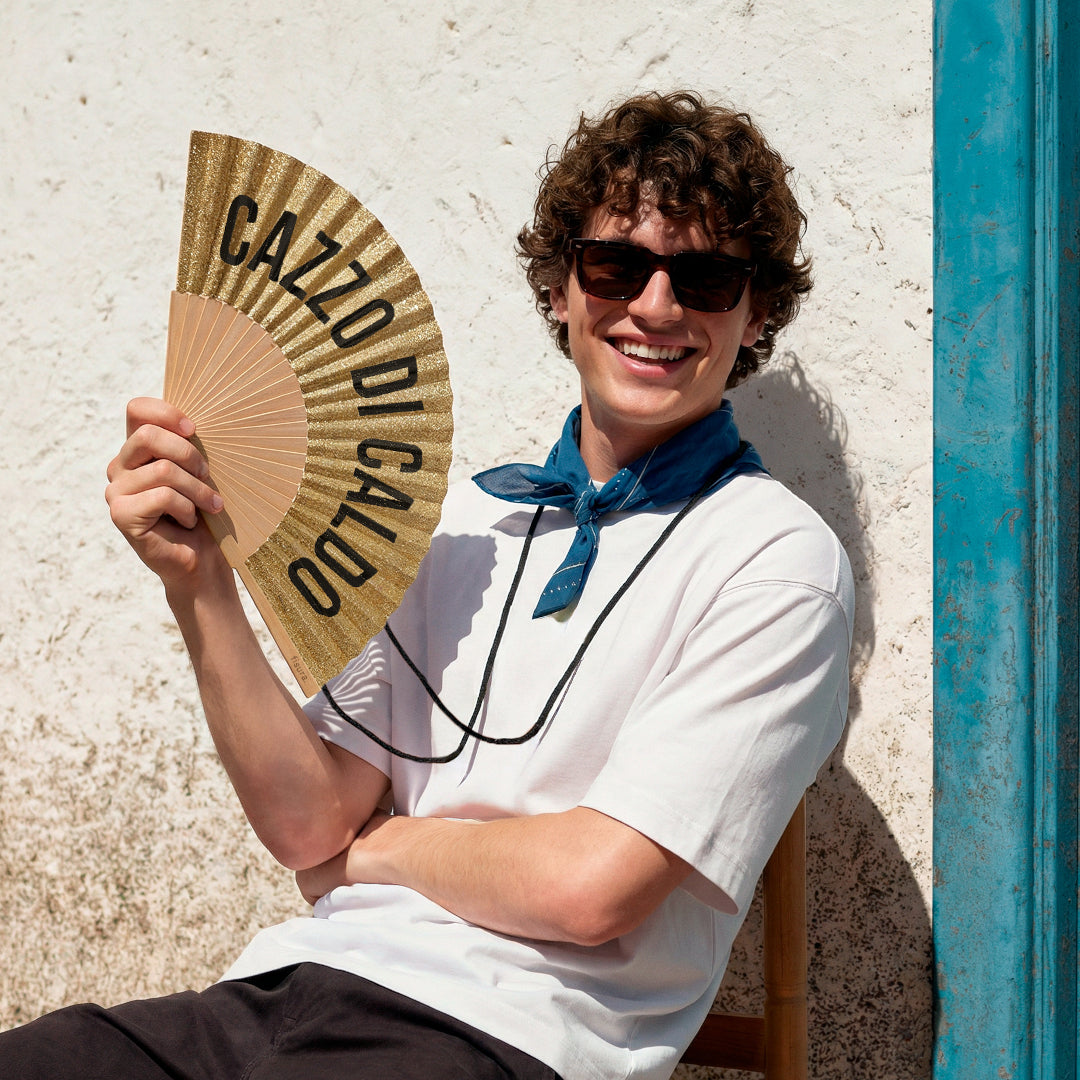 chico abanicándose con el abanico de la marca fisura de tela y madera en color purpurina dorada con el texto CAZZO DI CALDO en negro, con la cuerda negra colgada al cuello y sentado en una terraza al sol
