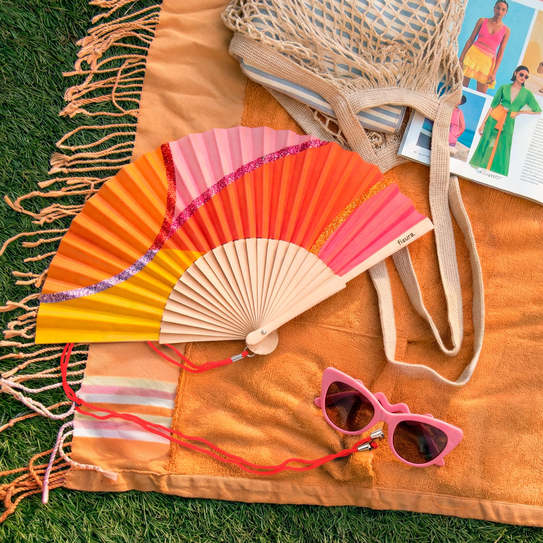 abanico de la marca fisura con ondas en colores amarillo, naranja y rosa, y con purpurina de colores,, ocn la cuerda para colgarlo en color rojo, sobre una toalla naranja en el césped con una bolsa y unas gafas de sol