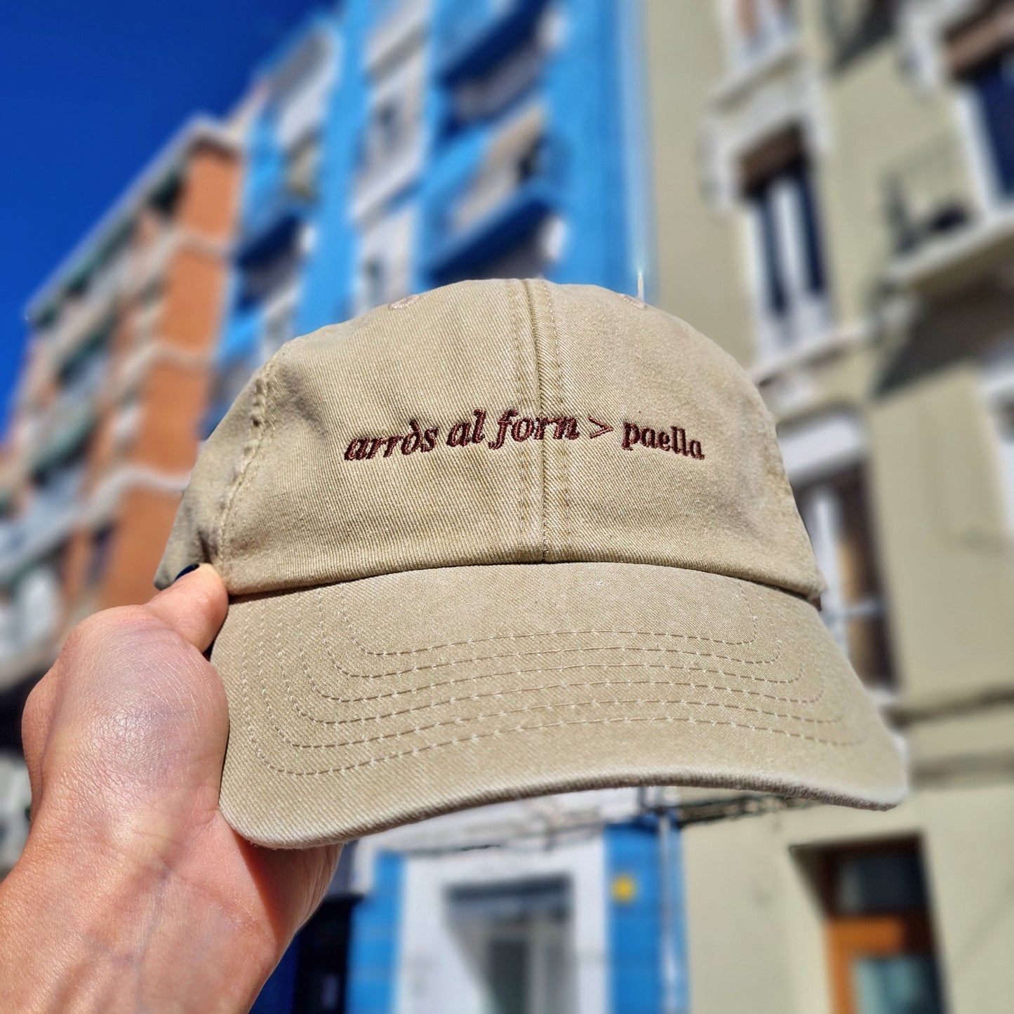 mano sujetando la gorra en color beige con el texto bordado en marrón que pone arròs al forn>paella
