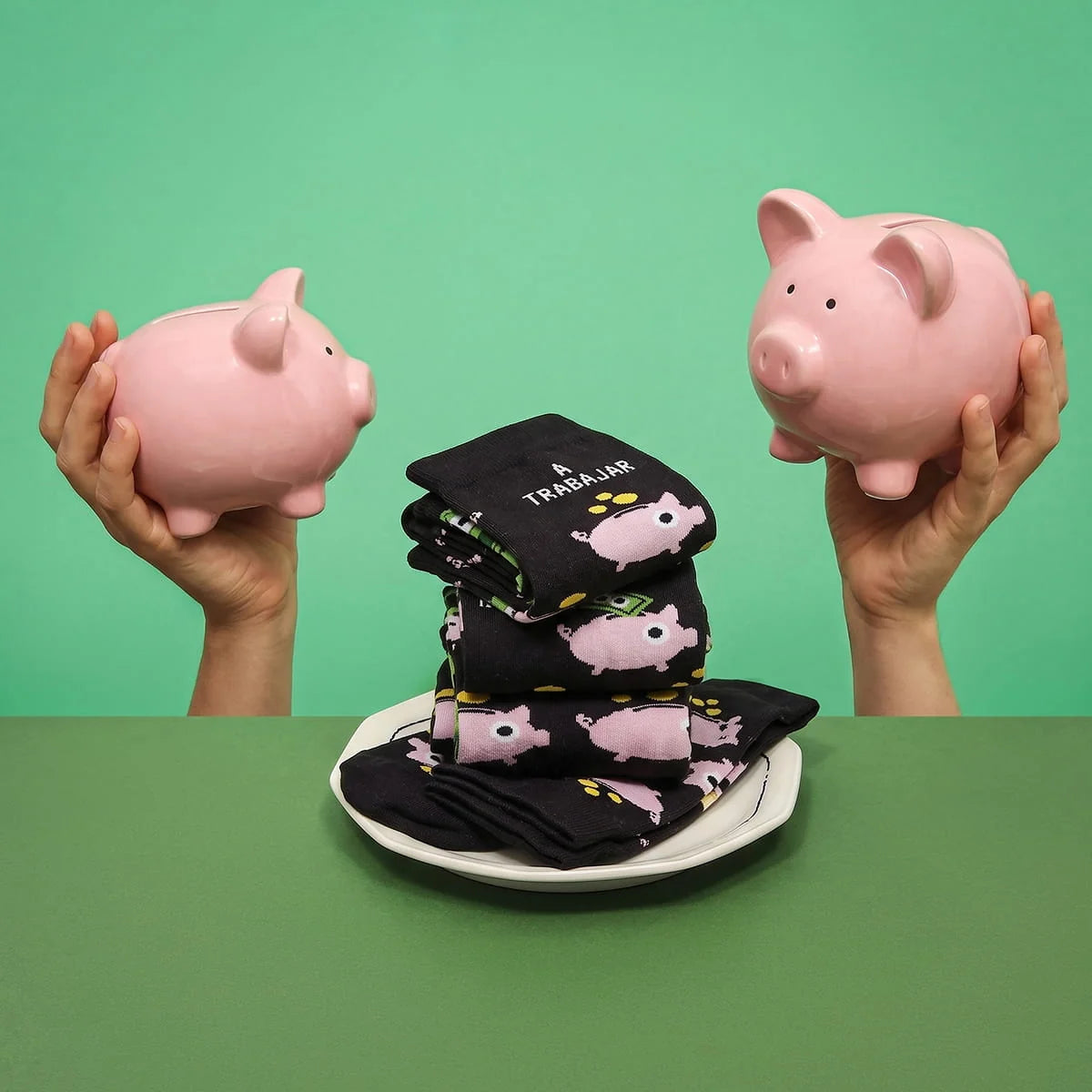 Calcetines UO «A trabajar» doblados sobre un plato, entre dos huchas rosas sostenidas por unas manos, sobre fondo verde.