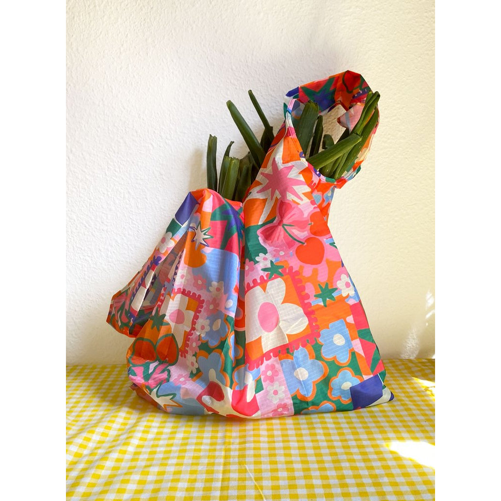 bolsa reusable de Laura Ortiz en colores rojo, verde, naranja, rosa y azul sobre un mantel a cuadros y con puerros dentro