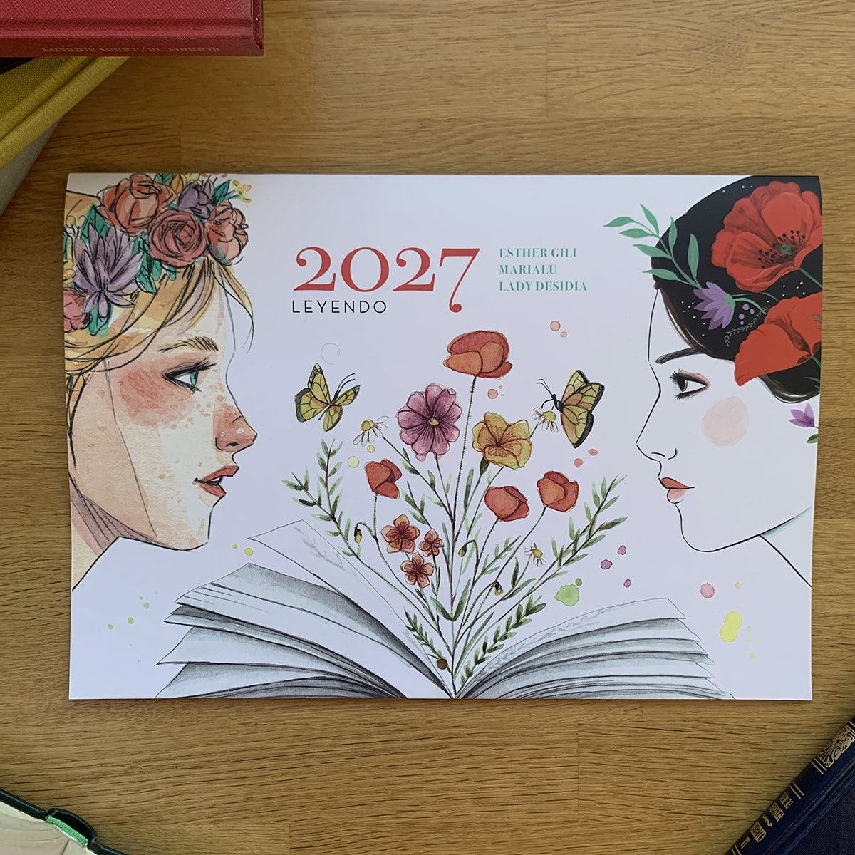 Portada del calendario 2027 Leyendo de Esther Gili, Marialu y Lady Desidia, con un libro abierto, flores y dos rostros femeninos.
