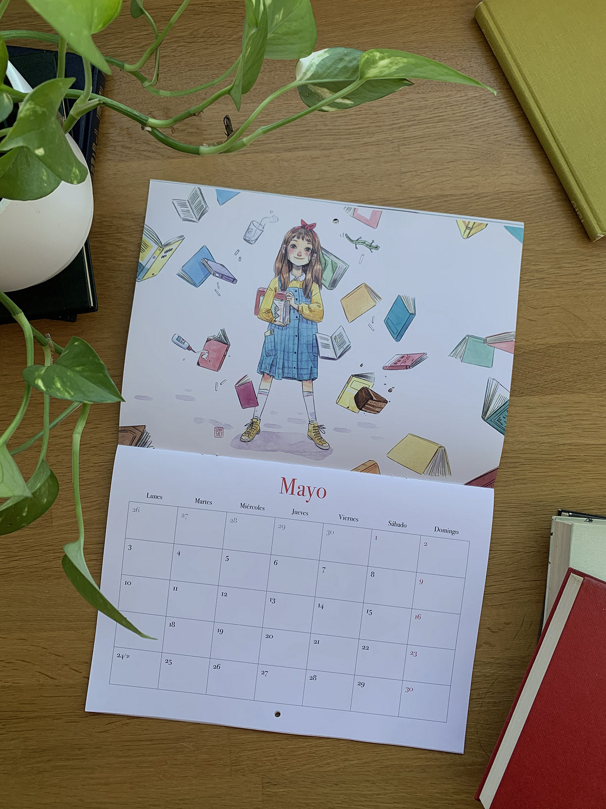 Calendario 2027 Leyendo abierto en mayo, con una niña rodeada de libros flotantes.