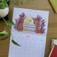 Calendario 2027 Leyendo abierto en julio, con una joven apoyada sobre una pila de libros entre flores moradas.