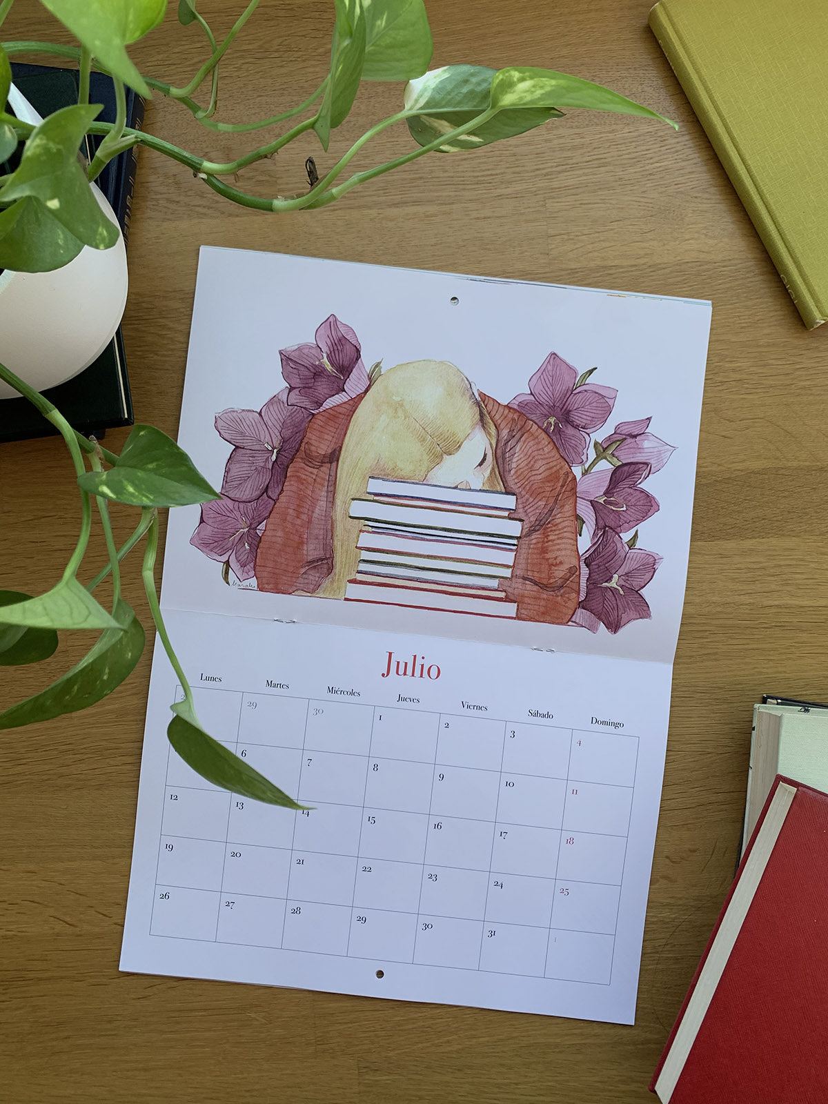 Calendario 2027 Leyendo abierto en julio, con una joven apoyada sobre una pila de libros entre flores moradas.