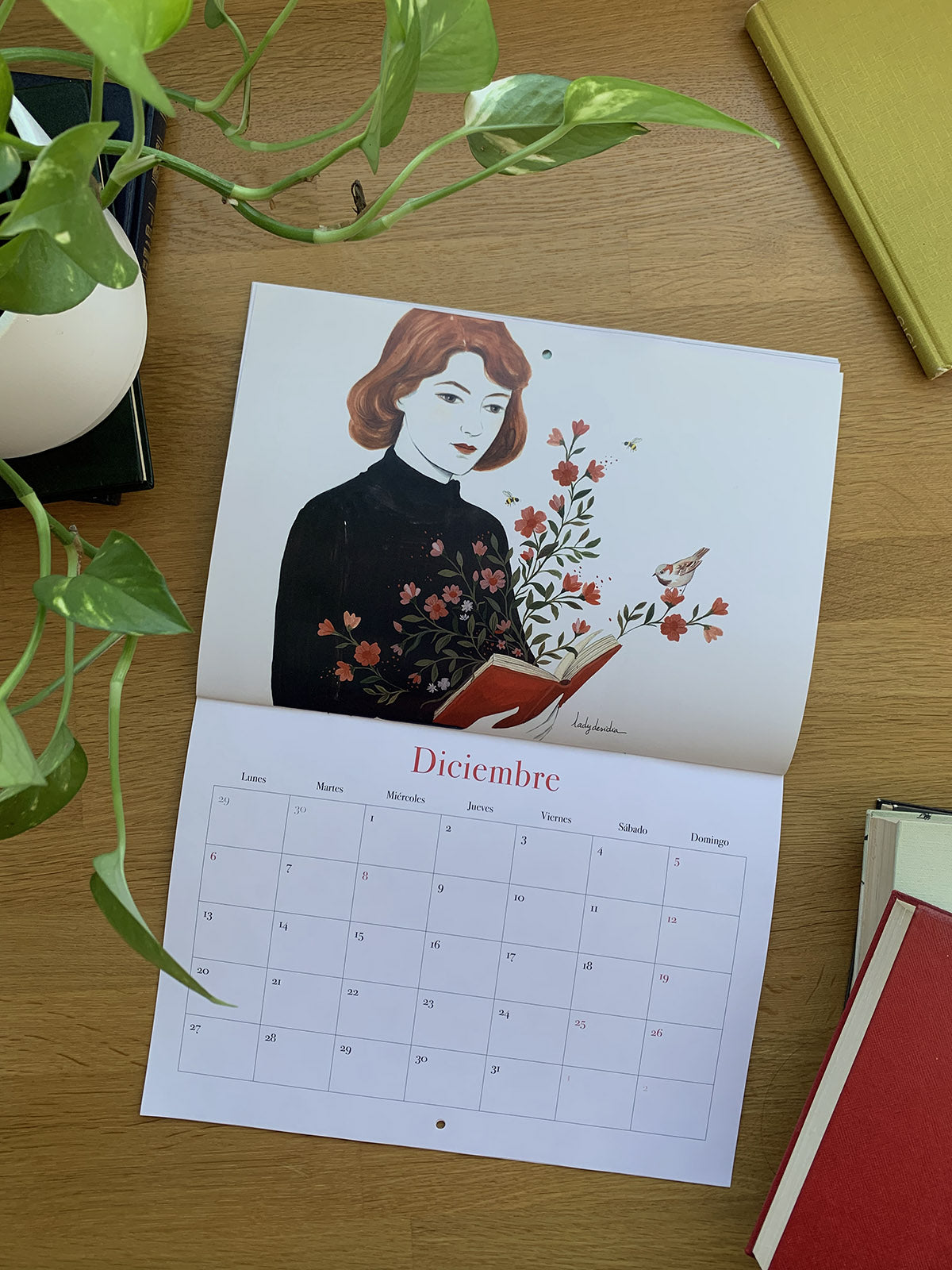 Calendario 2027 Leyendo abierto en diciembre, con una mujer sosteniendo un libro del que brotan flores.