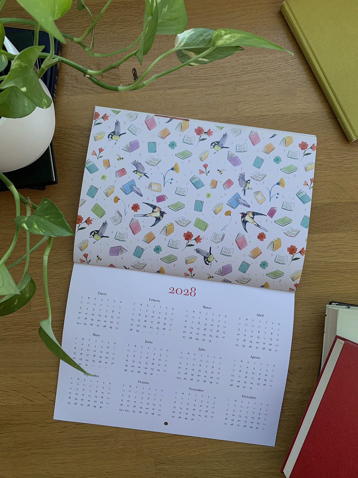 Calendario 2027 Leyendo abierto en la vista anual de 2028, con una ilustración de libros, golondrinas y flores.