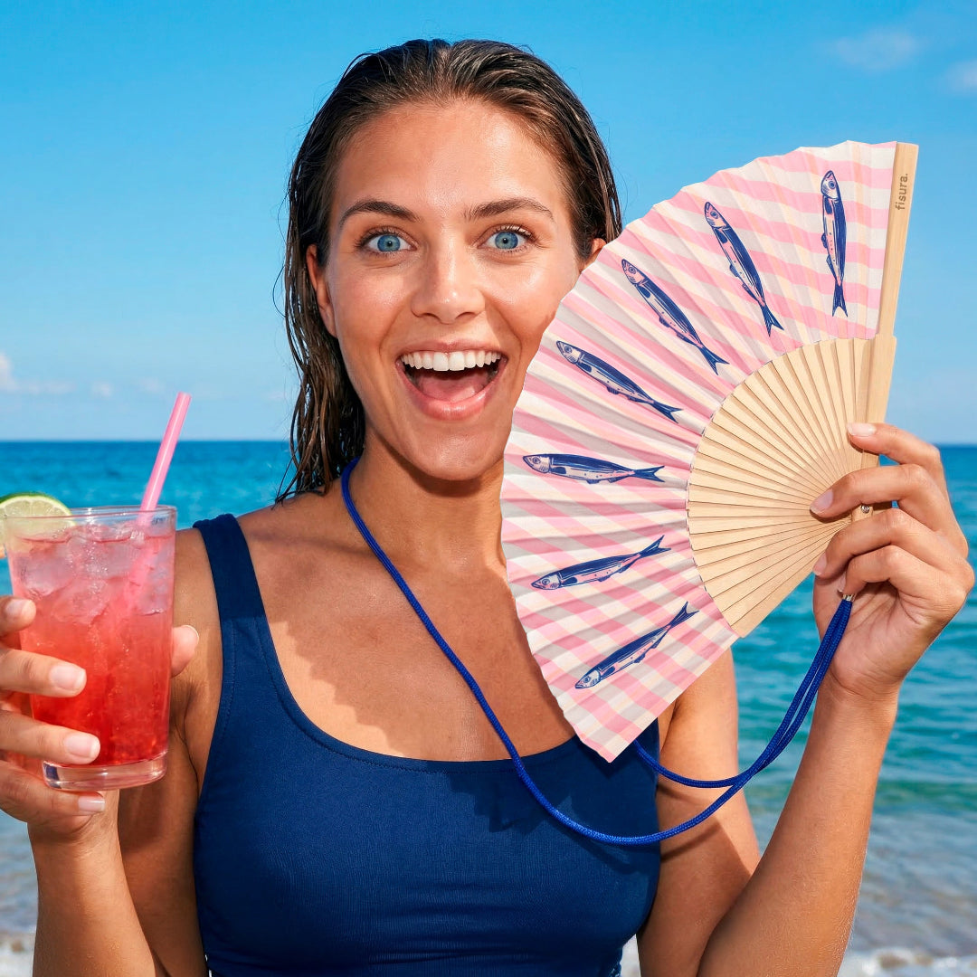 chica posando con el abanico de la marca fisura con el fondo a rayas verticales rosas y blancas y con estampado de sardinas en color azul oscuro y la cuerda en azul oscuro para colgarla al cuello, en el mar