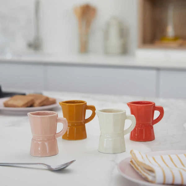 Juego de cuatro tazas de café expreso solo de cerámica de diferentes colores: naranja, blanco, caramel y rosa palo, en una mesa de cocina, junto a una cucharilla de café y platos