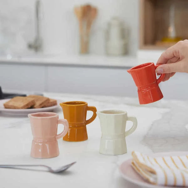 Juego de cuatro tazas de café expreso solo de cerámica de diferentes colores: naranja, blanco, caramel y rosa palo, en una mesa de cocina, junto a una cucharilla de café y platos. Con una mano sosteniendo la taza naranja