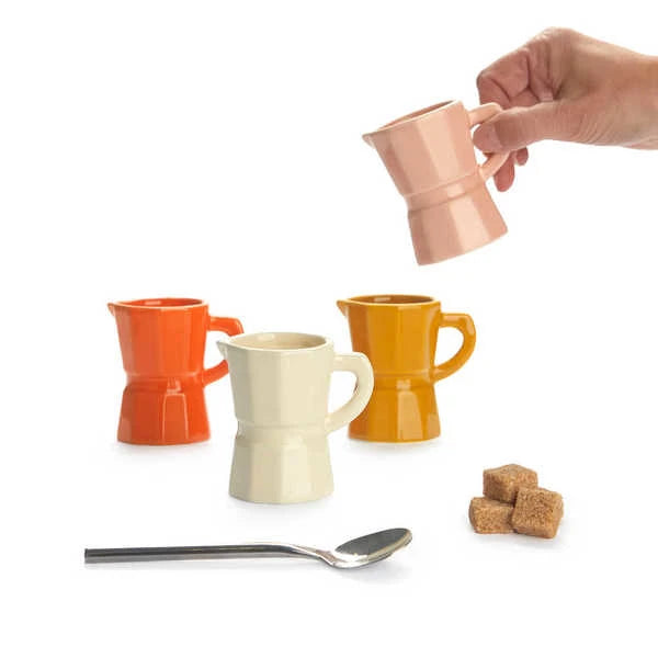 Juego de cuatro tazas de café expreso solo de cerámica de diferentes colores: naranja, blanco, caramel y rosa palo, sobre fondo blanco y con una mano sosteniendo la taza naranja. Al lado de una cuchara de café y unos terrones de azúcar