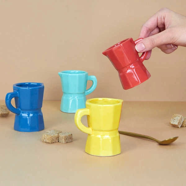 Juego de cuatro tazas de café expreso solo de cerámica de diferentes colores: azul, rosa, amarillo y turquesa junto a terrones de azúcar moreno y una cucharilla de café y terrones de azúcar, con una mano levantando la taza roja