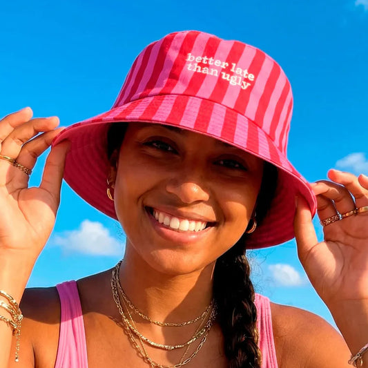 modelo con el gorro de Fisura reversible en color rosa a rayas y el texto better late than ugly en color blanco
