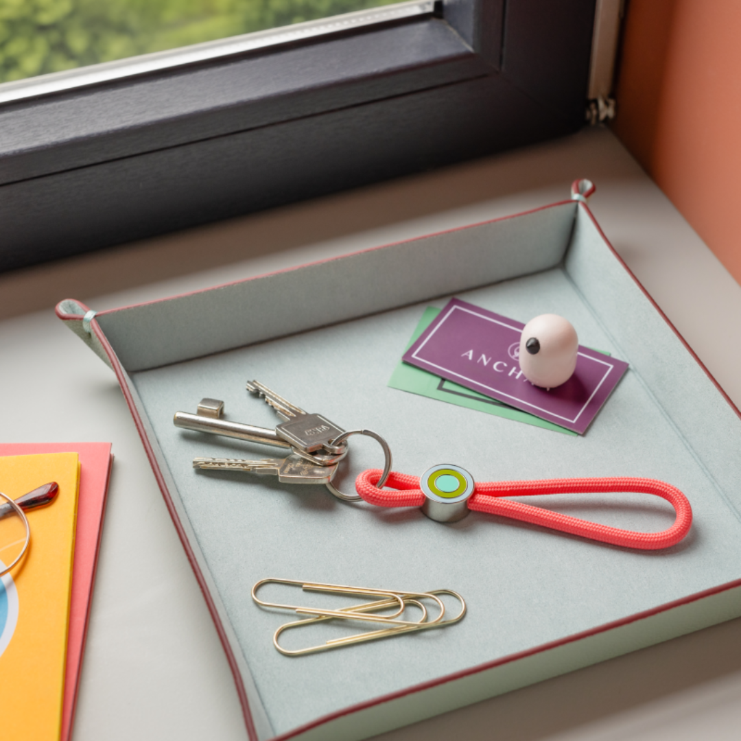 llavero colgante de cuerda roja para el móvil o las llaves con anilla plateada y un enganche con círculos de colores en un vacíabolsillos con clips y unas trajetas al lado