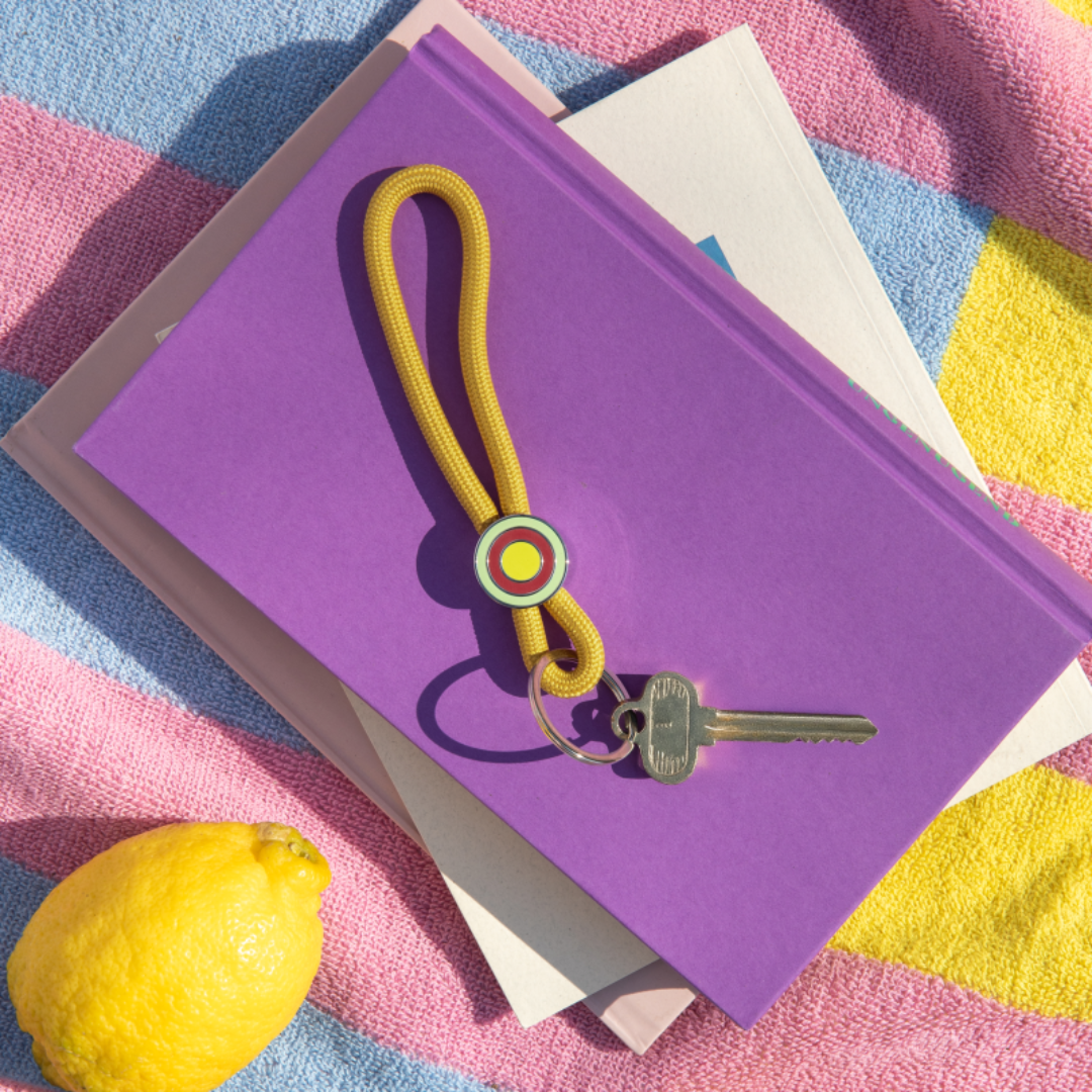 llavero colgante de cuerda amarillo mostaza para el móvil o las llaves con anilla plateada y un enganche con círculos de colores con una llave enganchada, sobre un libro morado con un limón al lado