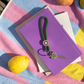 llavero colgante de cuerda verde oscuro para el móvil o las llaves con anilla plateada y un enganche con círculos de colores, con una llave colgada y sobre un libro y una toalla con un limón al lado