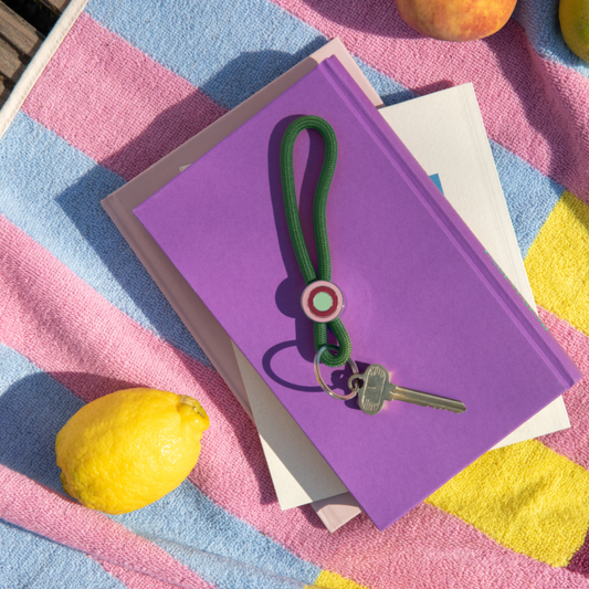 llavero colgante de cuerda verde oscuro para el móvil o las llaves con anilla plateada y un enganche con círculos de colores, con una llave colgada y sobre un libro y una toalla con un limón al lado