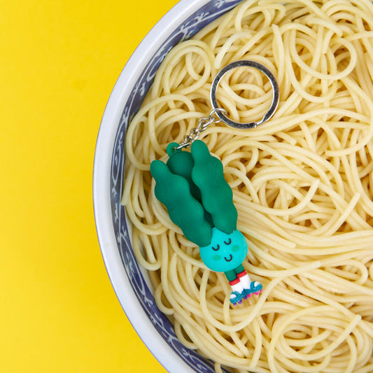 Llavero con forma de Pak Choi en color verde, con cara sonriente, calcetines blancos y patines reposando sobre un bol blanco y azul de fideos sobre fondo amarillo. 