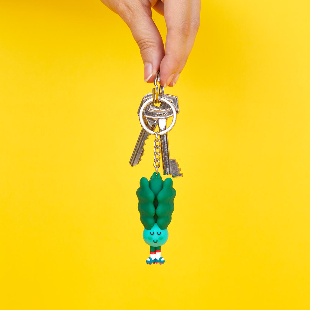 Llavero con forma de Pak Choi en color verde, con cara sonriente, calcetines blancos y patines cogido por una mano sobre fondo amarillo. 