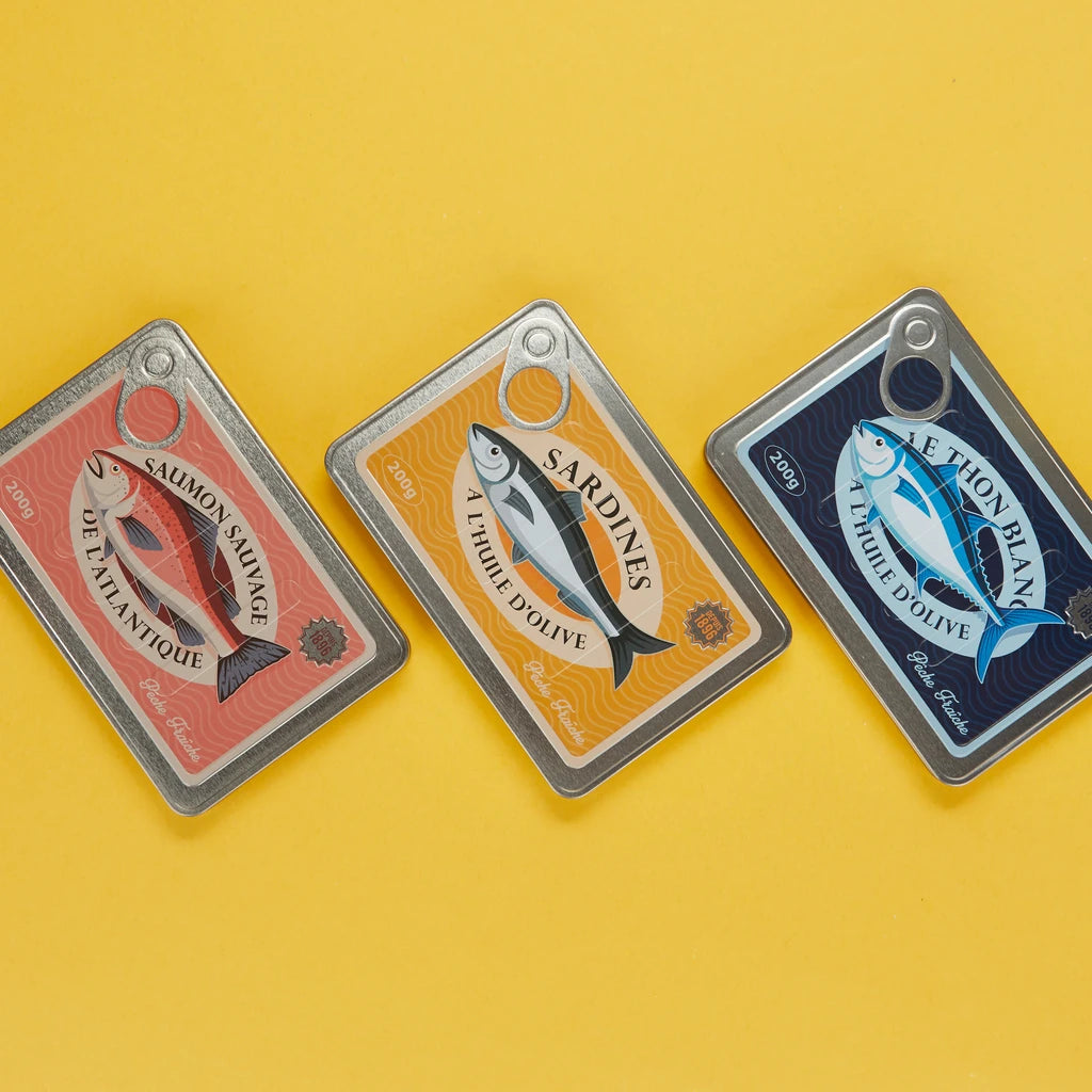conjunto de los tres modelos de libretas en forma de lata de conservas, en color rojo con un salmón, amarillo con una sardina y azul con un atún. Sobre fondo amarillo