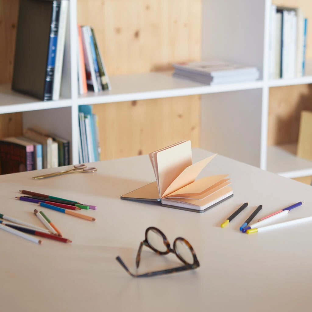 libreta en forma de lata de conservas en color rojo salmón abierta sobre una mesa con lápices y unas gafas, con las páginas teñidas en color salmón