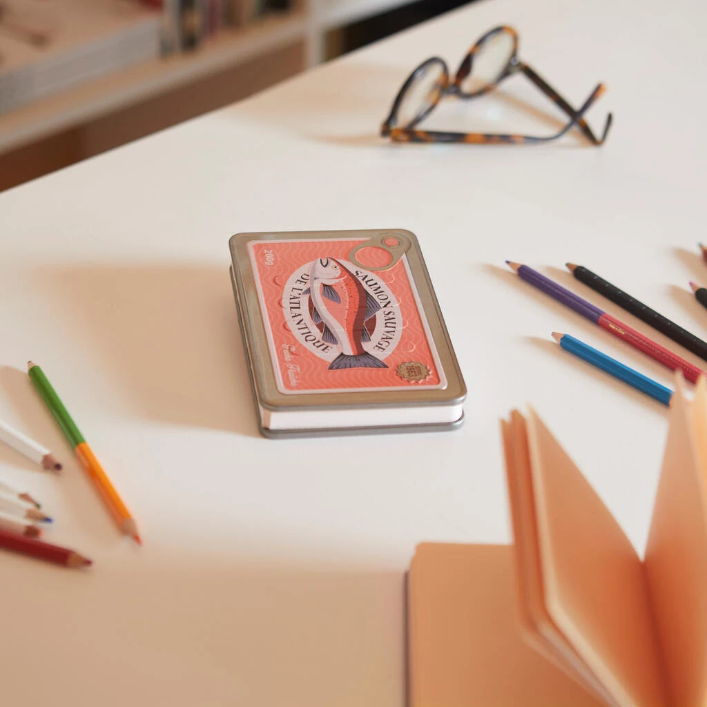 libreta en forma de lata de conservas en color rojo y con partes metálicas y un salmón en el centro con el texto Saumon sauvage de l'atlantique, cerrada sobre una mesa con lápices y unas gafas de vista