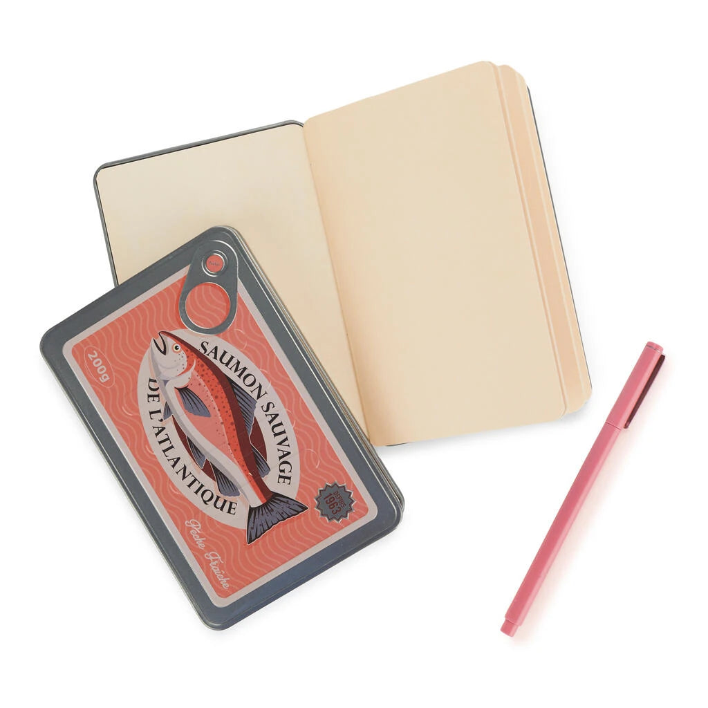 libreta en forma de lata de conservas en color rojo y con partes metálicas y un salmón en el centro con el texto Saumon sauvage de l'atlantique, la libreta abierta y un boli de color rosa