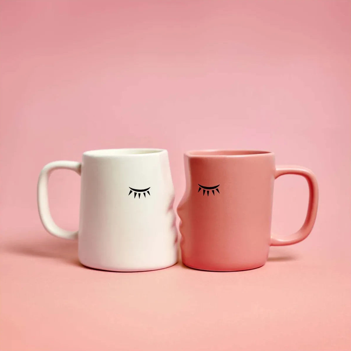 set de dos tazas en forma de caras de perfil que encajan dándose un beso con los ojos cerrados, una en color blanco y otra en color rosa. Sobre fondo rosa