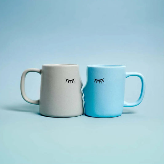 set de dos tazas en forma de caras de perfil que encajan dándose un beso con los ojos cerrados, una en color gris y otra en color azul. Sobre fondo azul