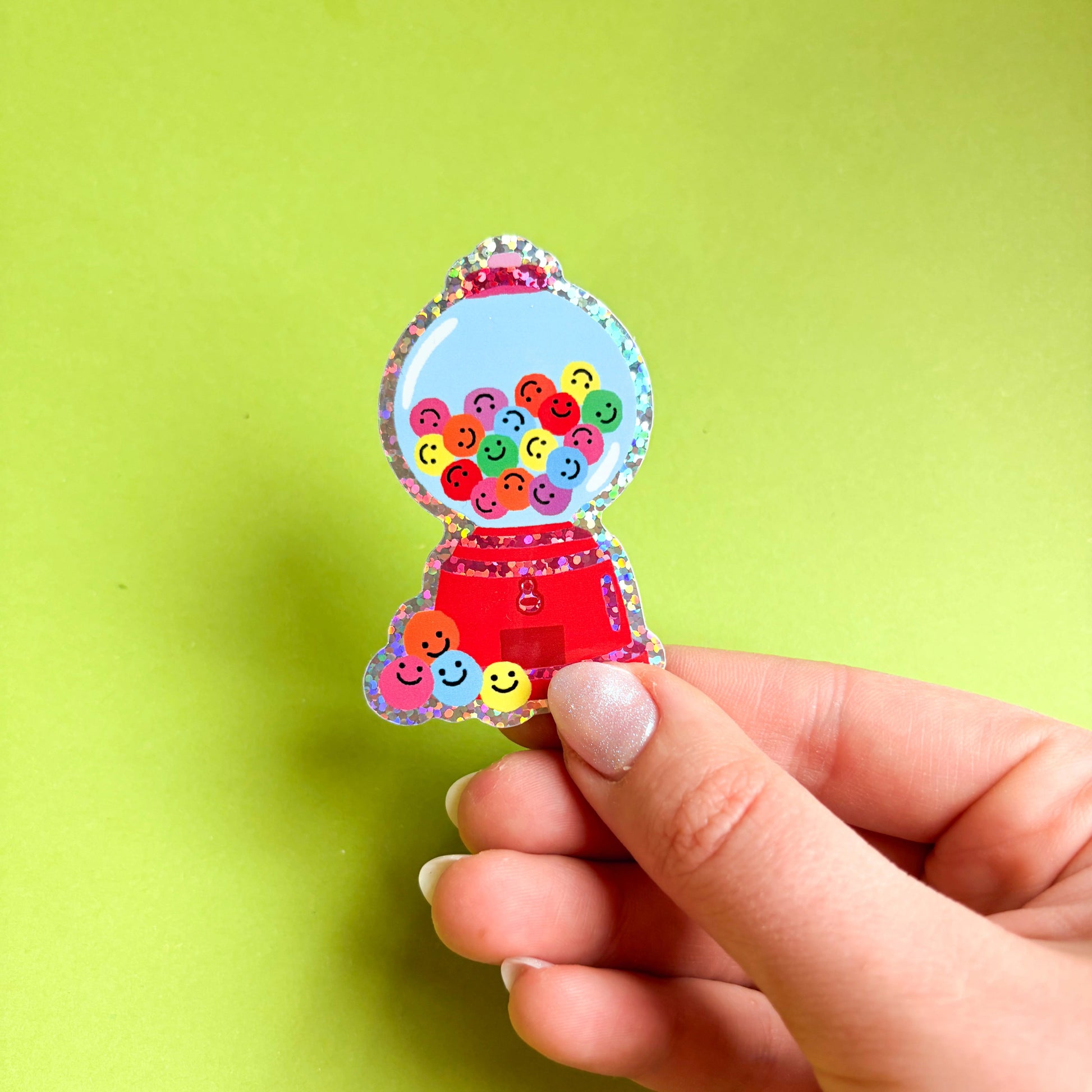 Mano sosteniendo una pegatina con brillo de una máquina expendedora de chicles roja, con bolas de colores con caritas sonrientes, sobre fondo verde claro.