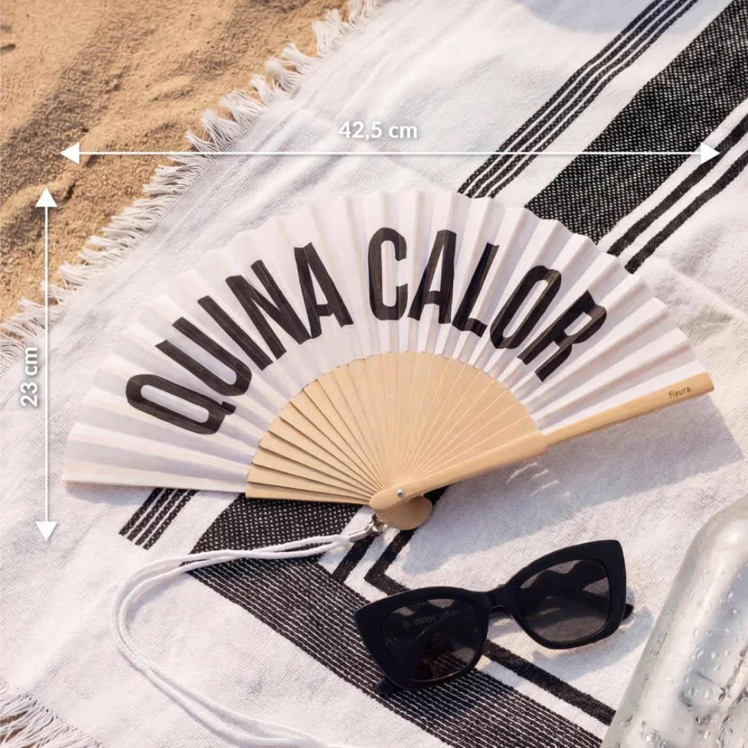 abanico de fisura en color blanco de tela y madera con el texto en negro "QUINA CALOR" abierto y con las medidas, sobre una toalla de playa al lado de unas gafas de sol