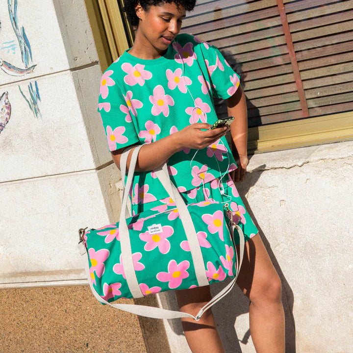modelo posando con la bolsa de viaje Simon de la marca Hindbag en colaboración con We are Jolies en color verde con patrón de flores rosas con el centro amarillo y las asas beige con un conjunto de camiseta y pantalón con el mismo patrón 