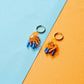 pendientes de metacrilato con arete dorado de la artista mitumi de unas sillas una encima de la otra, una en color naranja translúcido y la otra en color azul sólido
