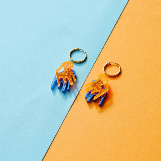 pendientes de metacrilato con arete dorado de la artista mitumi de unas sillas una encima de la otra, una en color naranja translúcido y la otra en color azul sólido