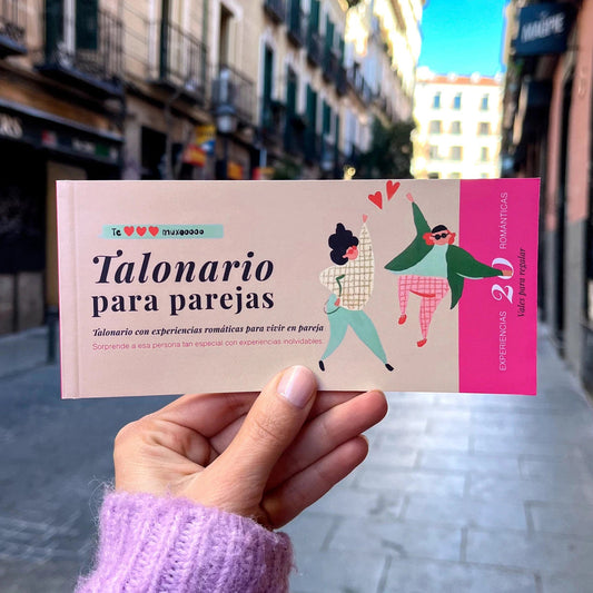 mano sujetando un talonario en color rosa con dos personas dibujadas saltando y el texto "talonario para parejas con experiencias románticas para vivir en pareja" con una ciudad de fondo.