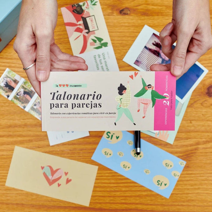 manos sujetando el talonario en color rosa con dos personas dibujadas saltando y el texto "talonario para parejas con experiencias románticas para vivir en pareja" con talones de fondo sobre una mesa