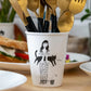 taza de porcelana con el dibujo en negro de una chica con el pelo largo con una planta a los pies y sujetando un gato negro y con cubiertos dentro de la taza