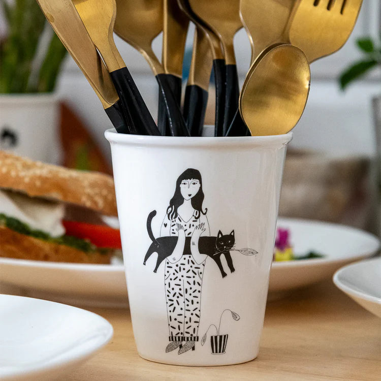 taza de porcelana con el dibujo en negro de una chica con el pelo largo con una planta a los pies y sujetando un gato negro y con cubiertos dentro de la taza
