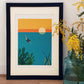 Un marco en color negro para fotos o prints con una lámina de Elisa Talens en la que se ve na mujer buceando en el mar está apoyado sobre una pared junto a un jarrón con flores amarillas.