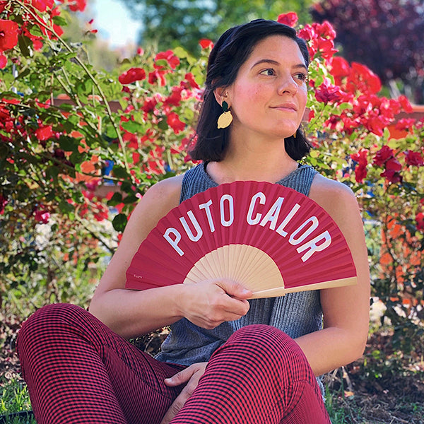 Mujer posando en verano con un abanico rojo con letras blancas sobre un fondo de flores y plantas