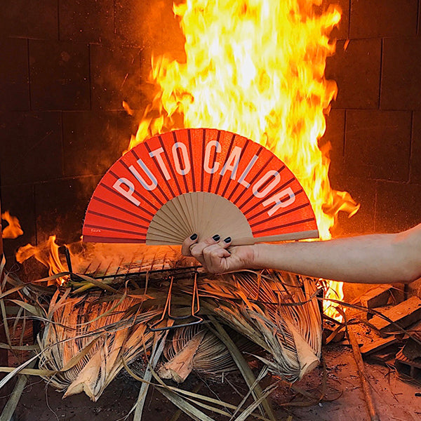 Mano sujetando un abanico rojo donde pone puto calor delante de una llama de fuego