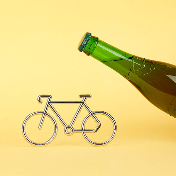 Botella de cerveza cerrada y abridor metálico con forma de bici sobre fondo amarillo 