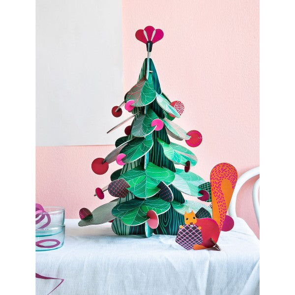 Árbol de Navidad montable de cartón de hojas verdes y con decoración de bolitas rojas y piñas y una figurita de ardilla sobre una mesa de fiesta