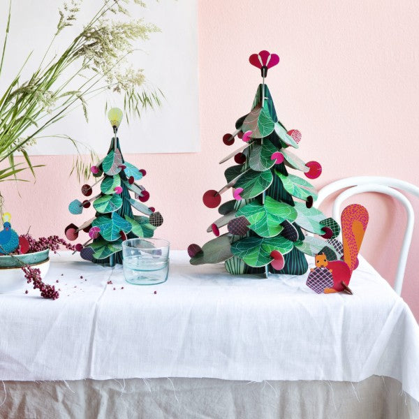 Dos árboles de Navidad montables de cartón de hojas verdes y con decoración de bolitas rojas y piñas y una figurita de pavo real
